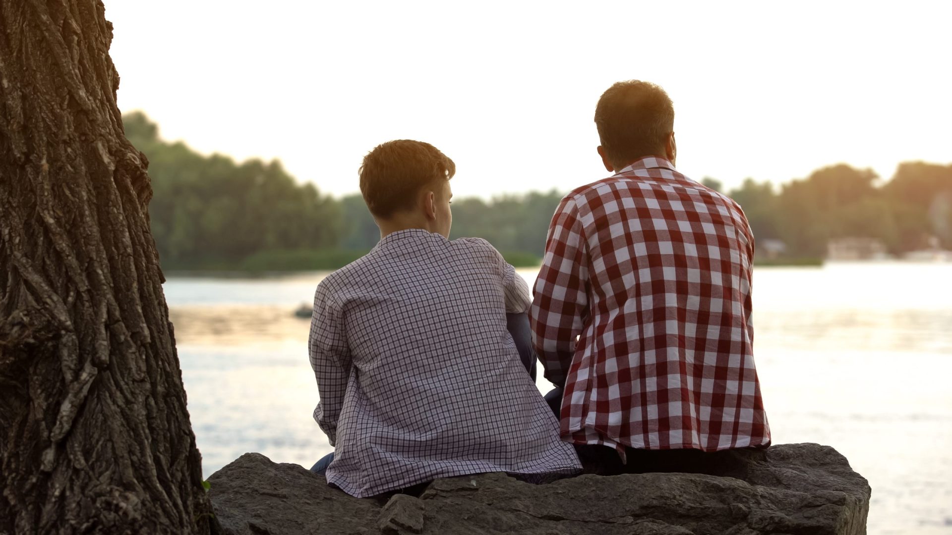 Male,Teenager,And,His,Father,Sitting,On,Stone,Near,Lake Parent and teen sitting facing away from the camera talking to each other