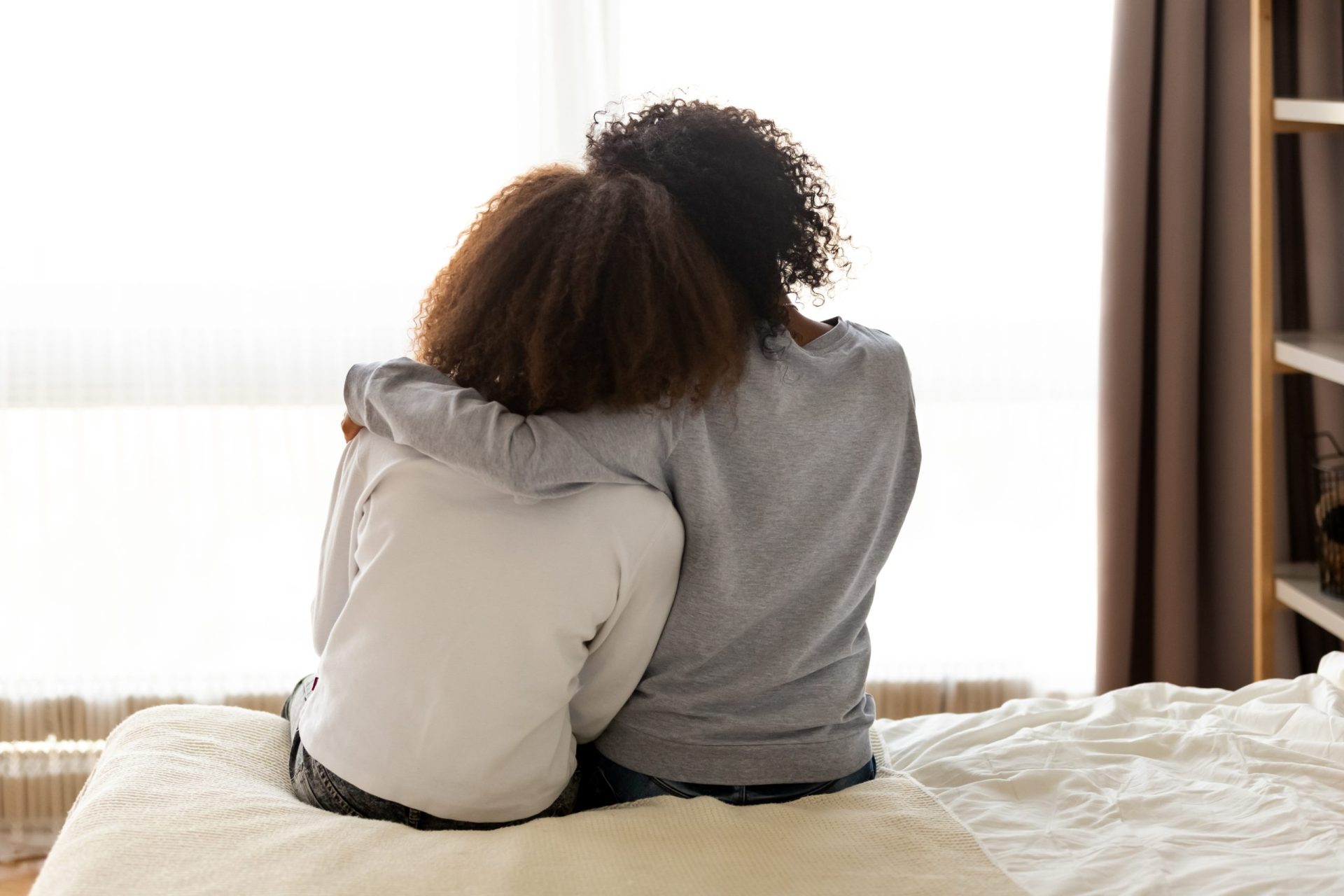Rear,Back,View,Black,Mother,And,Daughter,Embrace,Sitting,On two people sitting on a bed comforting one another