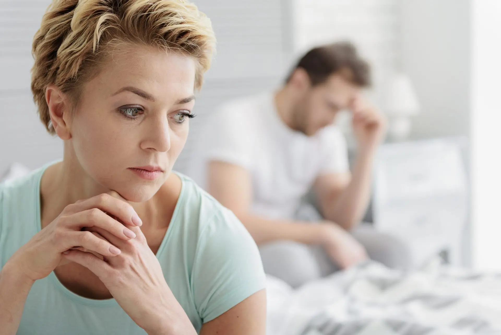 Portrait of pensive middle-aged woman thinking about her marriage with sadness. Her husband is sitting on bed