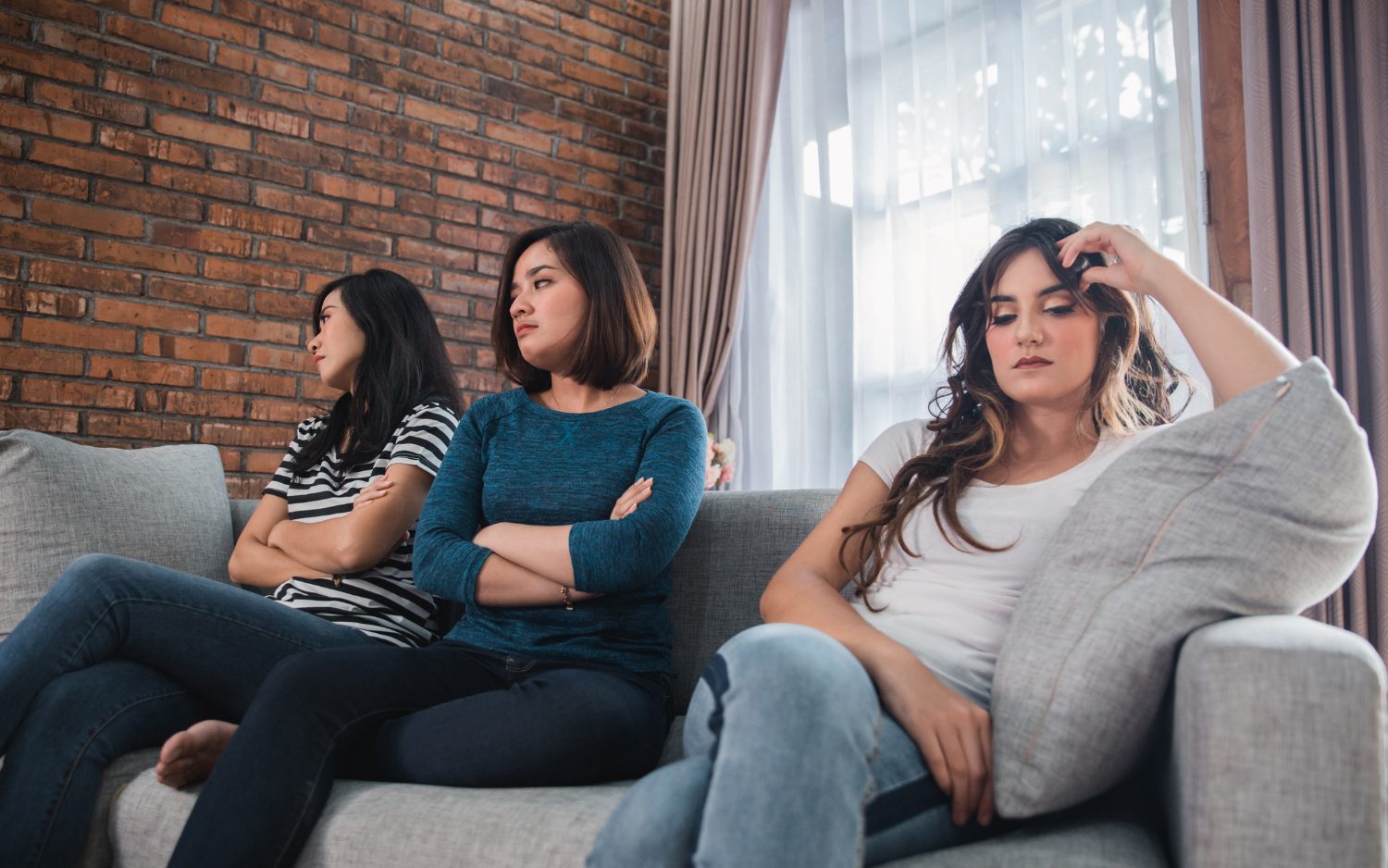 Group of three teenage girls sitting on a couch looking unhappy two sitting closer together one sitting farther away